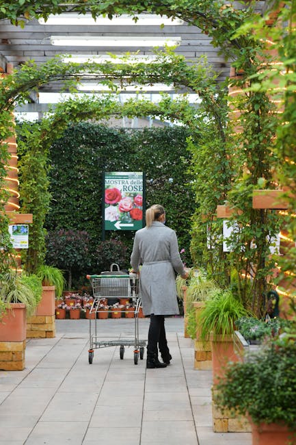 A woman with her hair tied back, dressed in a light gray coat and dark trousers, stands in a greenhouse or conservatory, pushing a metal shopping cart filled with small plant pots along a paved pathwa